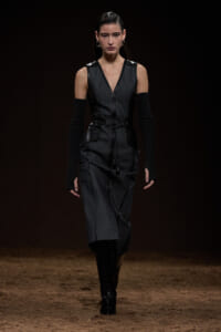 Model walking a runway in a sleeveless dark gray denim dress with a waist belt and long black knit arm warmers, paired with black knee-high boots on a dimly lit catwalk.