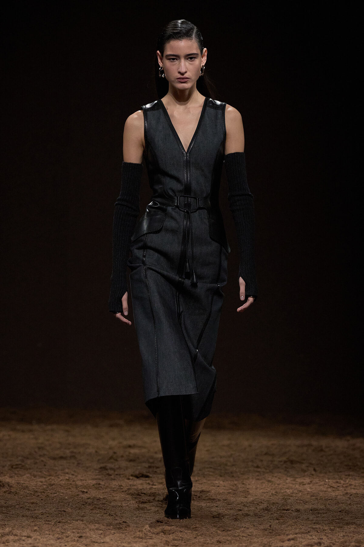Model walking a runway in a sleeveless dark gray denim dress with a waist belt and long black knit arm warmers, paired with black knee-high boots on a dimly lit catwalk.