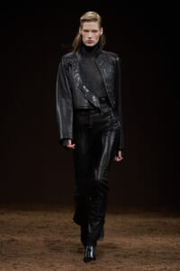 Model walking a fashion show in a black leather jacket and matching pants with quilted panels on the front.