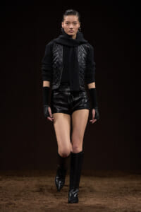 Model walking in a black, quilted vest over a chunky sweater and scarf, paired with black leather shorts and tall black boots on a runway.
