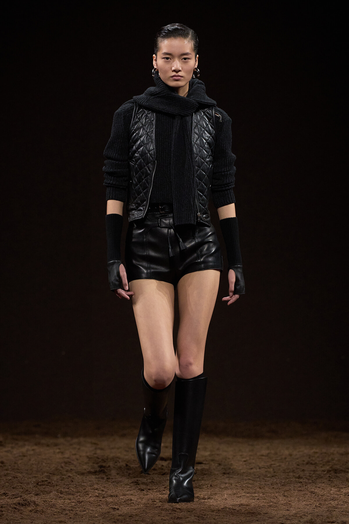 Model walking in a black, quilted vest over a chunky sweater and scarf, paired with black leather shorts and tall black boots on a runway.