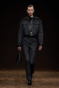 Model walking a runway in a dark denim outfit with black leather trim, carrying a black handbag.