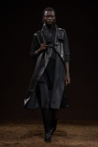 Model walking a runway in a long black leather sleeveless coat over a dark turtleneck and knee-high boots.