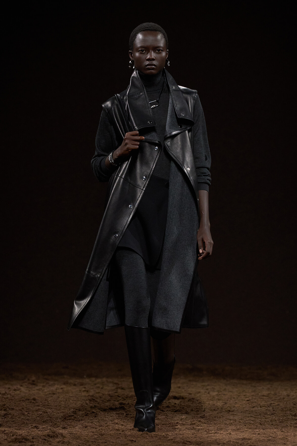 Model walking a runway in a long black leather sleeveless coat over a dark turtleneck and knee-high boots.