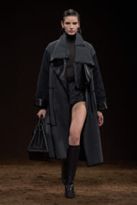 Model on a runway wearing a long gray coat with black leather panels, black turtleneck, knee-high boots, and a black structured handbag.
