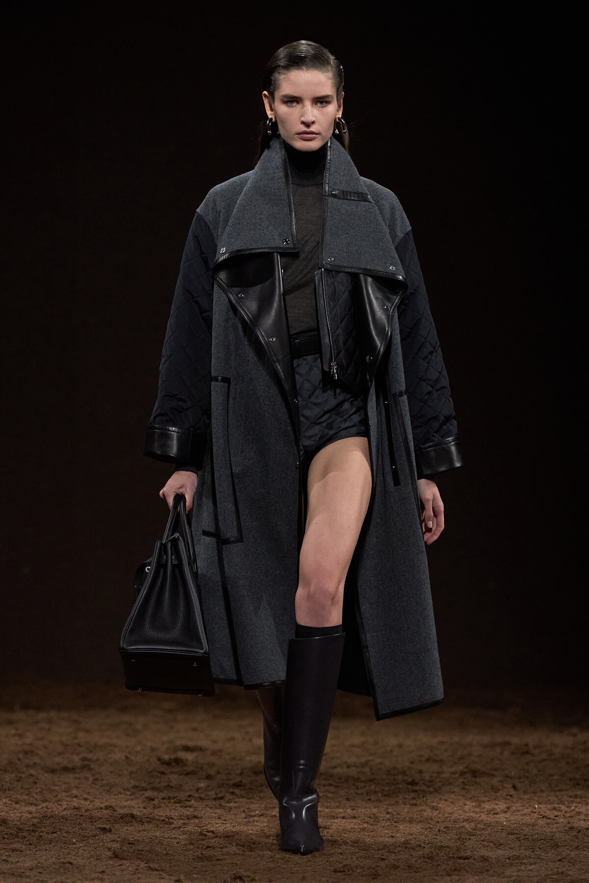 Model on a runway wearing a long gray coat with black leather panels, black turtleneck, knee-high boots, and a black structured handbag.
