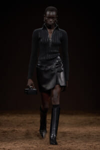 Black-clad model walking a fashion runway in a ribbed zip sweater, black leather mini wrap skirt, and knee-high boots, holding a small black box clutch.