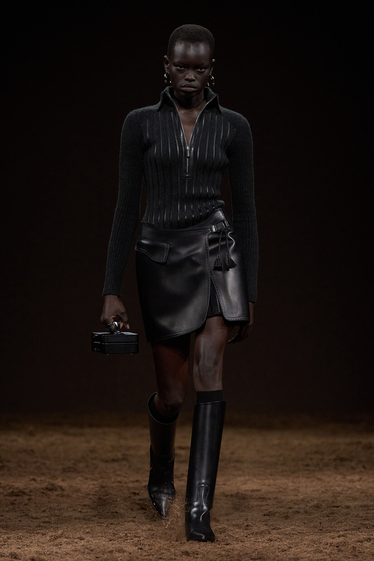Black-clad model walking a fashion runway in a ribbed zip sweater, black leather mini wrap skirt, and knee-high boots, holding a small black box clutch.