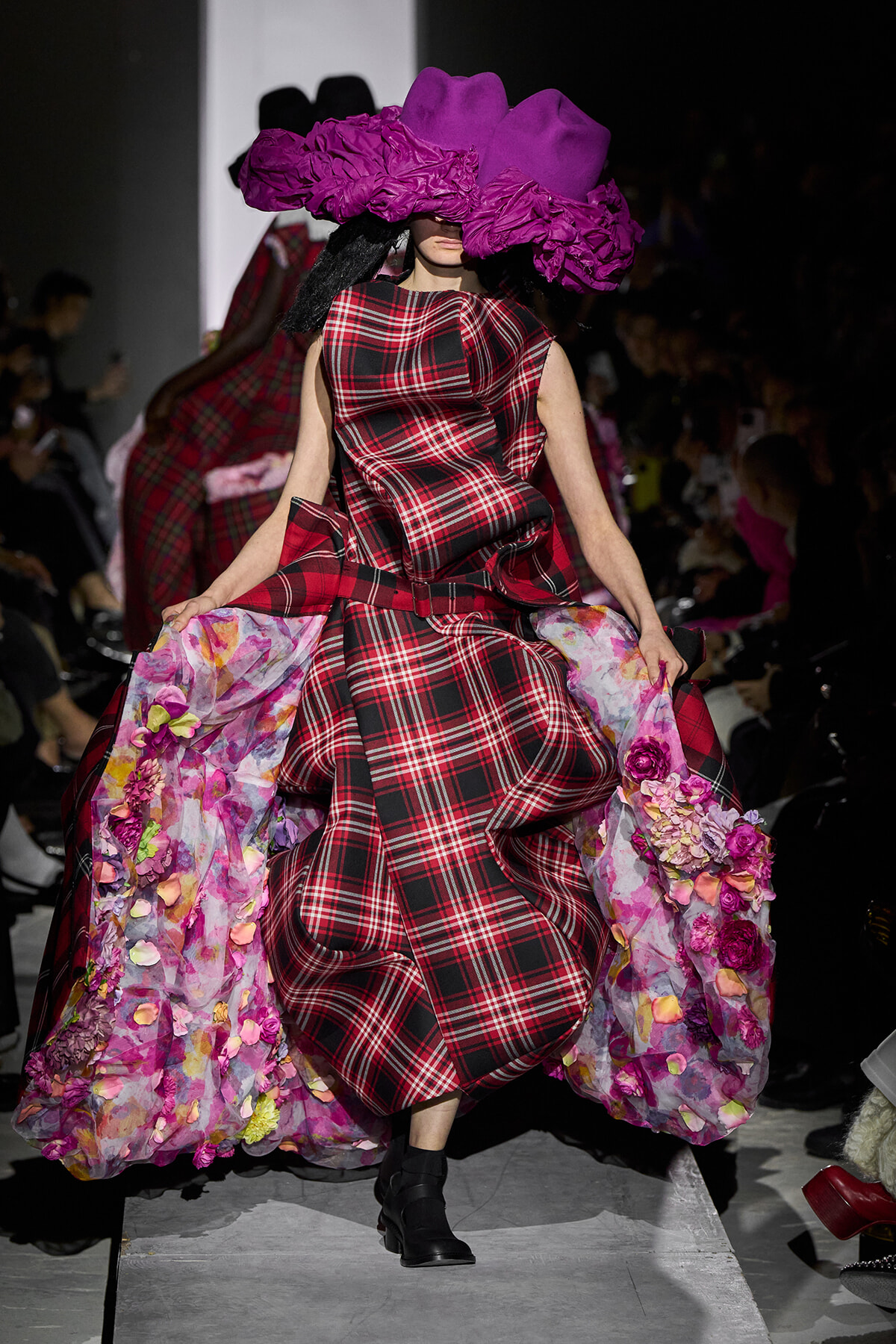 Model on runway wearing a red and black plaid gown with a voluminous pink floral lining and a large purple hat obscuring her face, mid-stride.