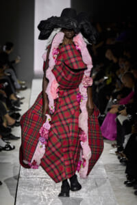 Model on a runway wearing a dramatic red plaid gown with pink ruffle trim and a large black hat