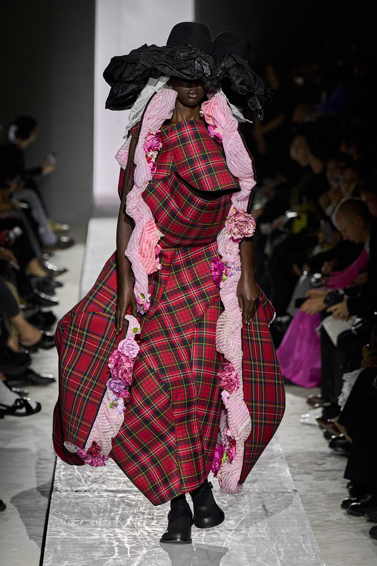 Model on a runway wearing a dramatic red plaid gown with pink ruffle trim and a large black hat