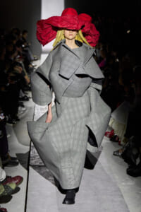 Model wearing an oversized gray plaid coat with sculptural sleeves on a runway, accented by a bold red hat obscuring her eyes.