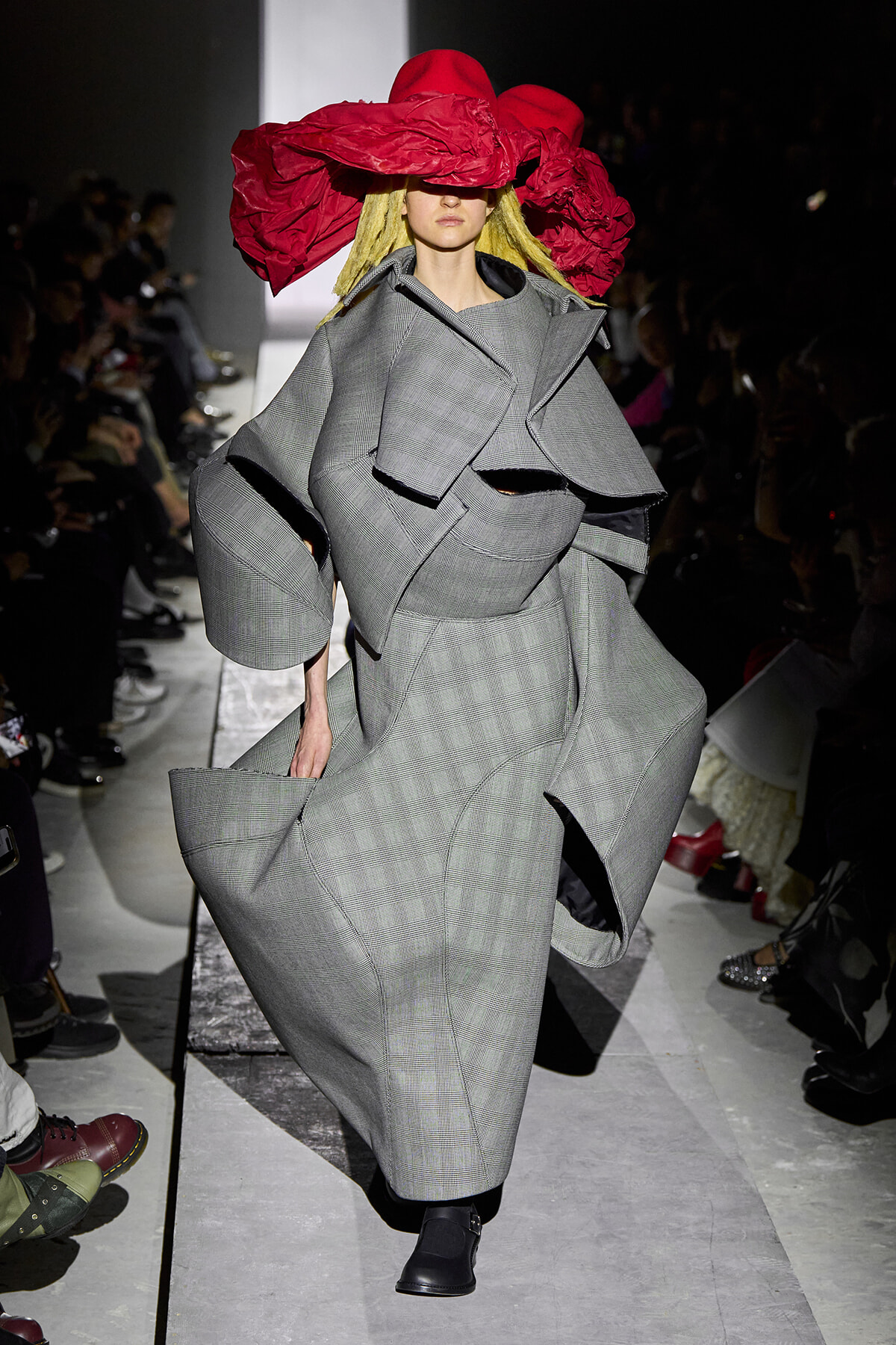 Model wearing an oversized gray plaid coat with sculptural sleeves on a runway, accented by a bold red hat obscuring her eyes.