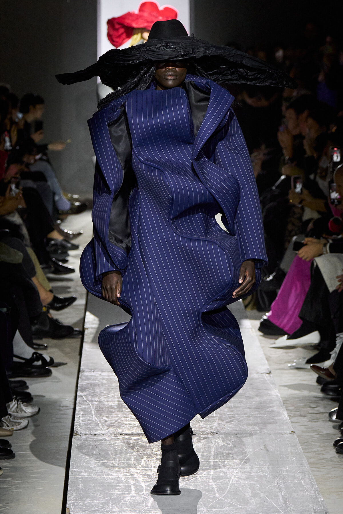 Model walks the runway in a structured blue pinstripe coat with oversized shoulders and a wide black hat, dramatic silhouette.