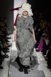 Model on a fashion runway wearing a black-and-white gingham dress with oversized ruffles and a large white sculptural headpiece.