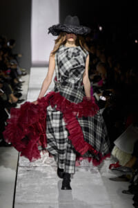 Model walks a checkered plaid gown with red ruffled train on a runway, wearing a wide black hat that obscures her eyes.