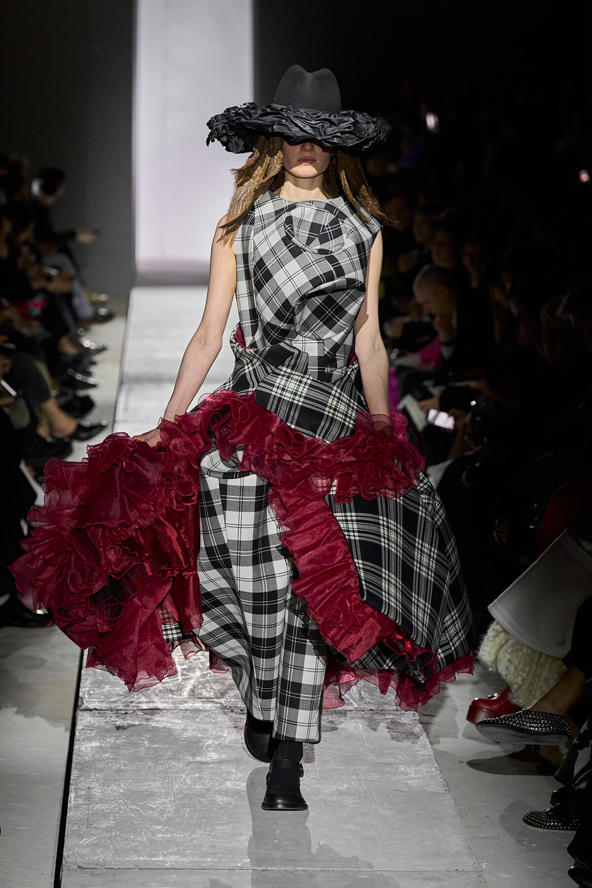Model walks a checkered plaid gown with red ruffled train on a runway, wearing a wide black hat that obscures her eyes.