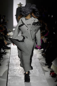 Model walks the runway in a black-and-white gingham suit with an oversized sculptural black hat.