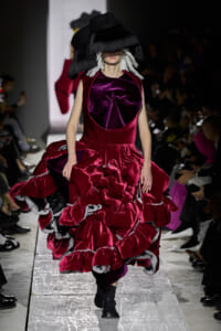 Model walks a fashion show in a burgundy velvet gown with a voluminous, ruffled skirt and a large black hat partially covering her face on a runway.