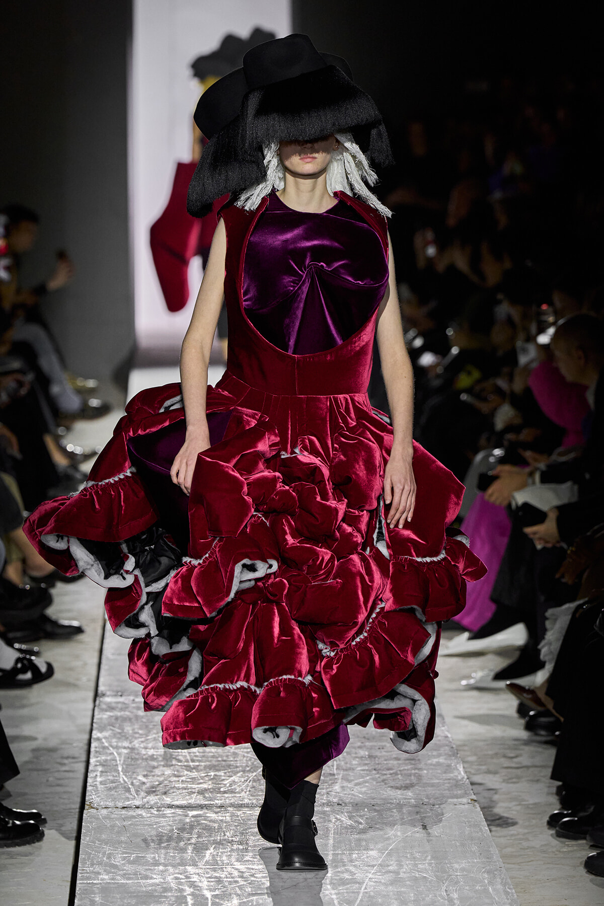 Model walks a fashion show in a burgundy velvet gown with a voluminous, ruffled skirt and a large black hat partially covering her face on a runway.