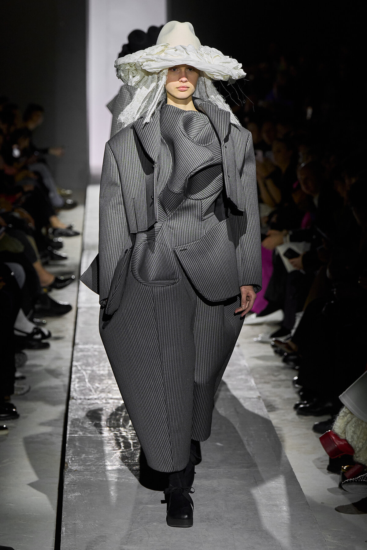 Runway model in a voluminous gray pinstripe suit with sculpted drapes and a large white hat, walking the catwalk beside a seated audience.