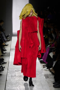 Model on a runway wearing a bold red pleated dress with a matching cape and a large yellow textured wig, walking toward the camera.