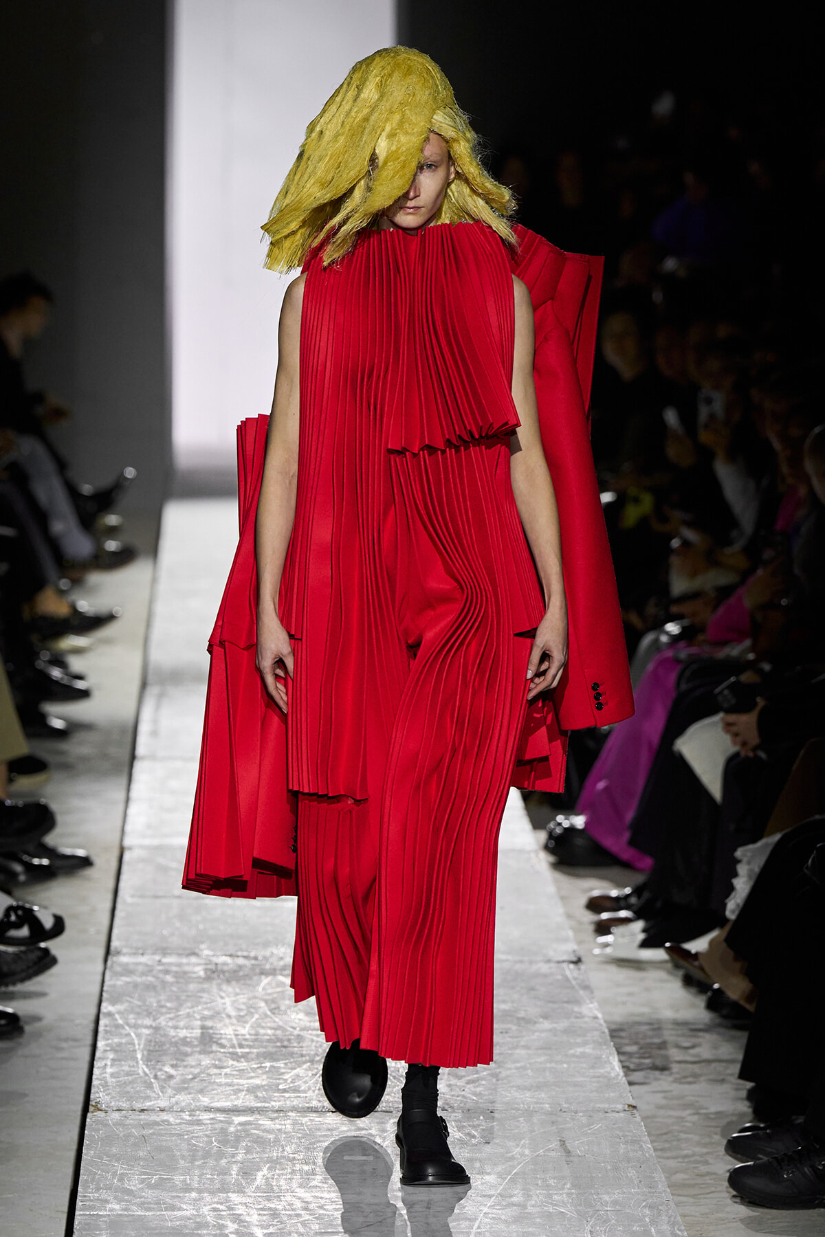 Model on a runway wearing a bold red pleated dress with a matching cape and a large yellow textured wig, walking toward the camera.