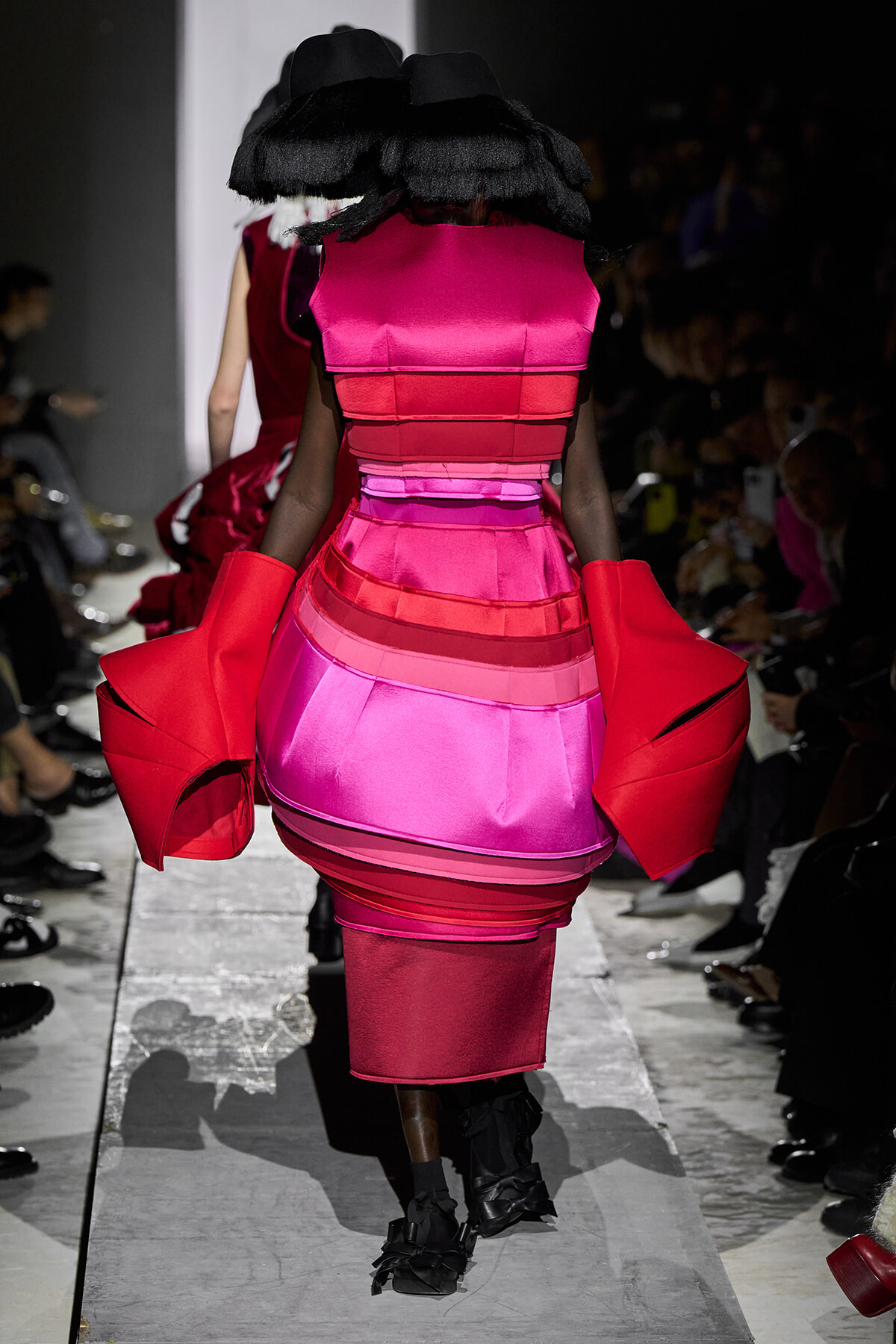 Back view of a model in a vibrant pink and red tiered dress with oversized red sleeves on a runway.