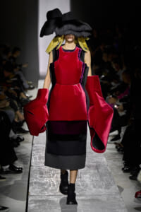 Model walks a runway in a bold red velvet dress with voluminous black lantern sleeves and color panels, paired with a large black hat obscuring her face.