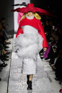 Model walks a fashion runway wearing a bright red cape over a white, ruffled gown and a large red hat that obscures the face.