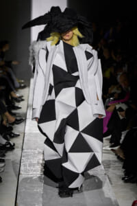 Model on a runway wearing an oversized black-and-white geometric coat with a large black hat.