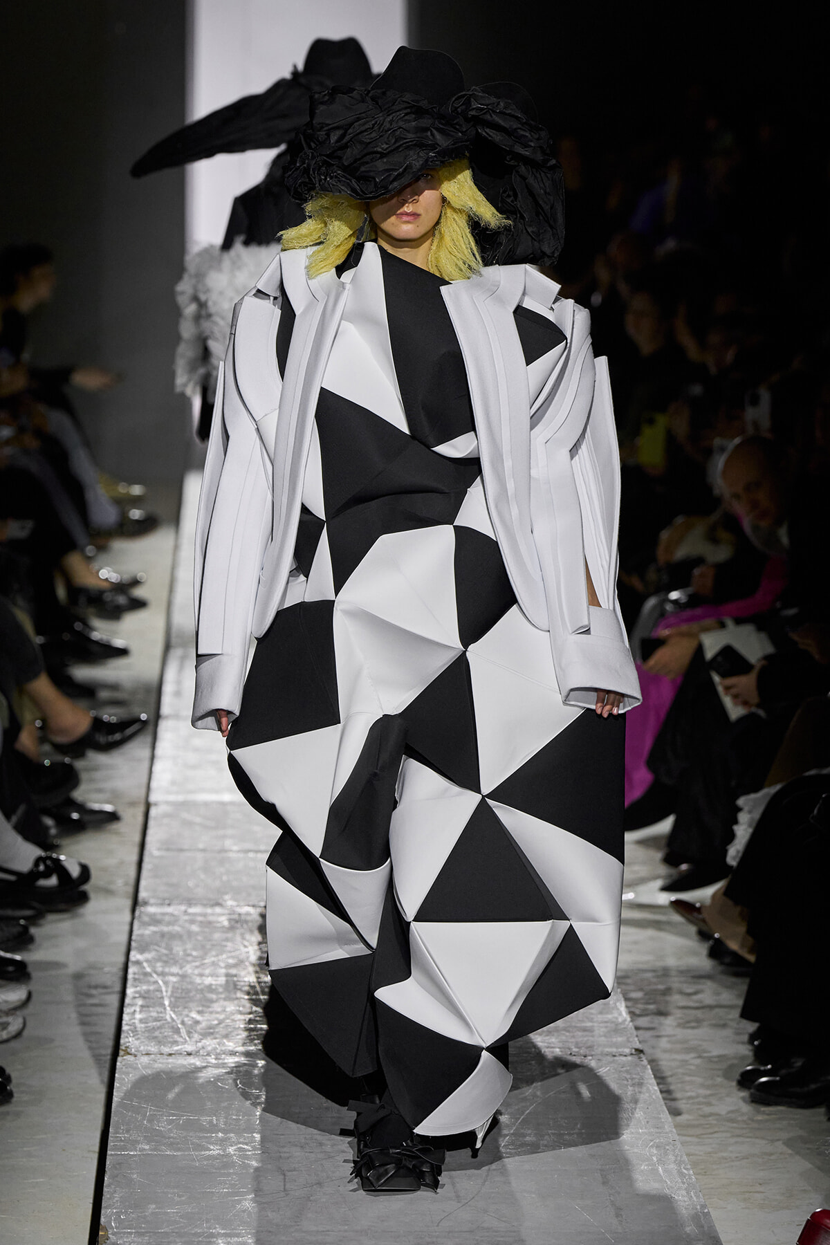 Model on a runway wearing an oversized black-and-white geometric coat with a large black hat.