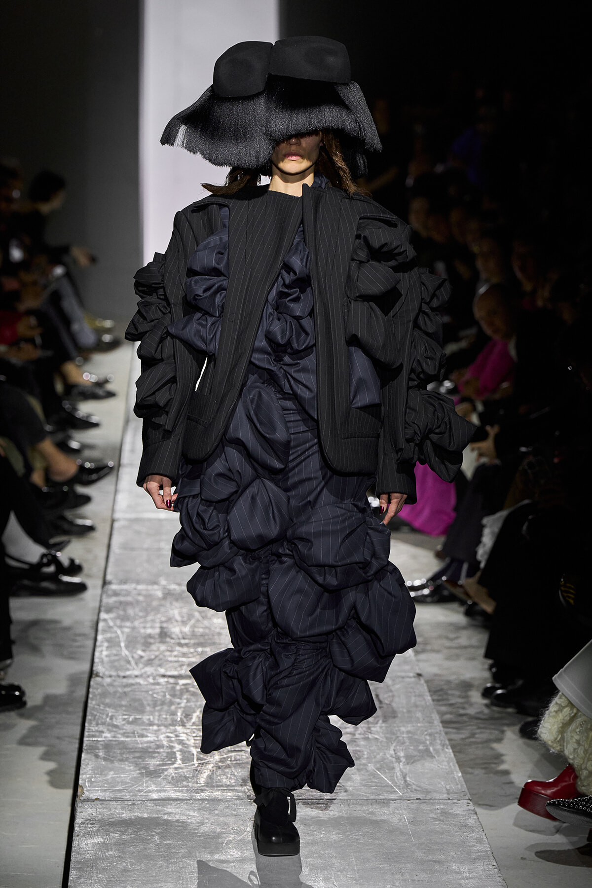 Model walks a dark, voluminous black gown with oversized ruffled layers and a wide brimmed hat that obscures her eyes on a fashion runway.