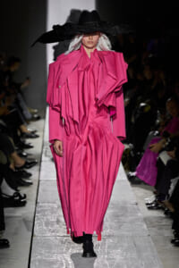 Model walks a fashion show wearing a vivid pink, oversized pleated gown with sculptural shoulders and a wide black hat on a runway.