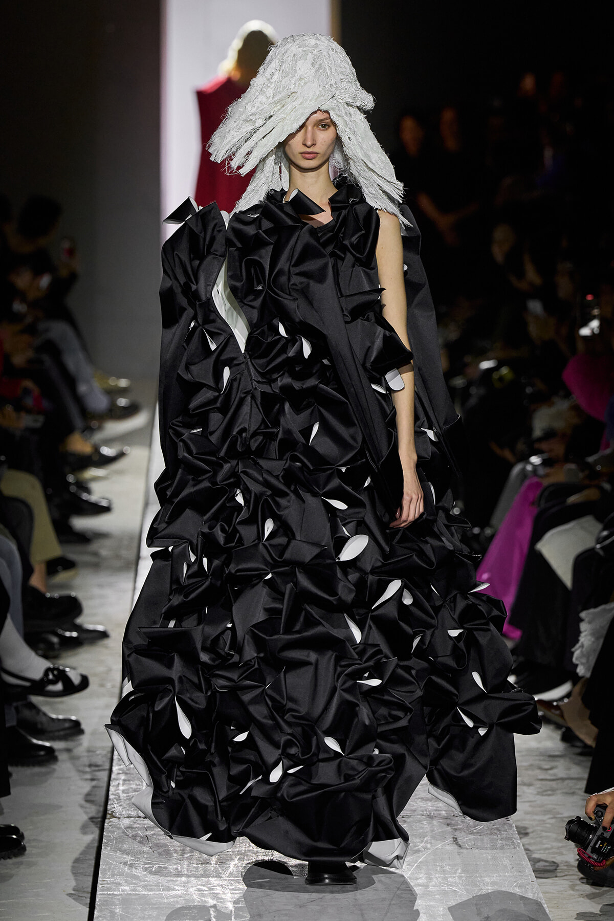 Model on a runway wearing a dramatic black sculptural gown with voluminous ruffles and white accents, paired with a textured white headpiece.