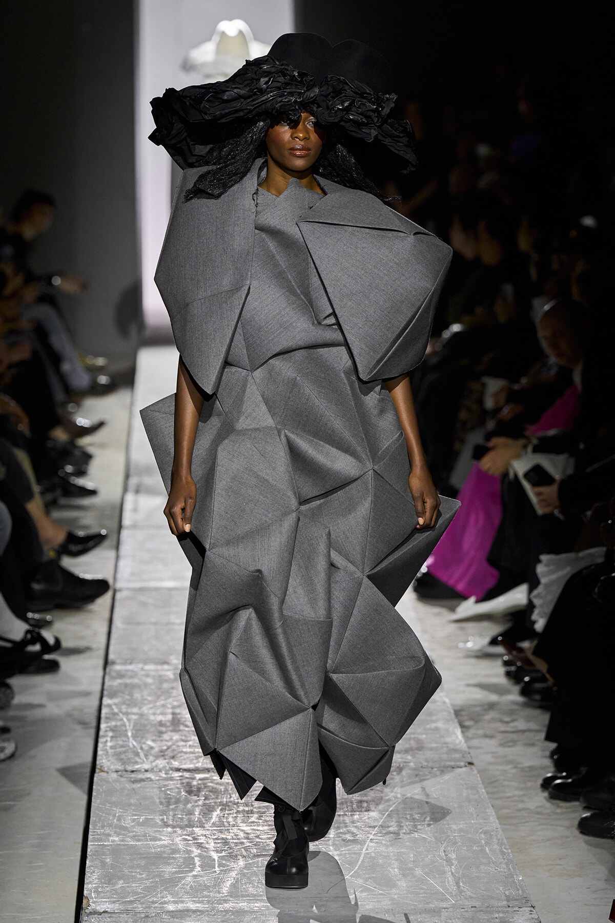 Model on a runway wearing a voluminous gray geometric dress and a large black ruffled hat, audience in background.