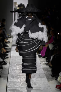 Runway model in a black pinstripe sculptural outfit with white ruffle sleeves and a wide-brimmed hat obscuring the face, black ankle boots visible.