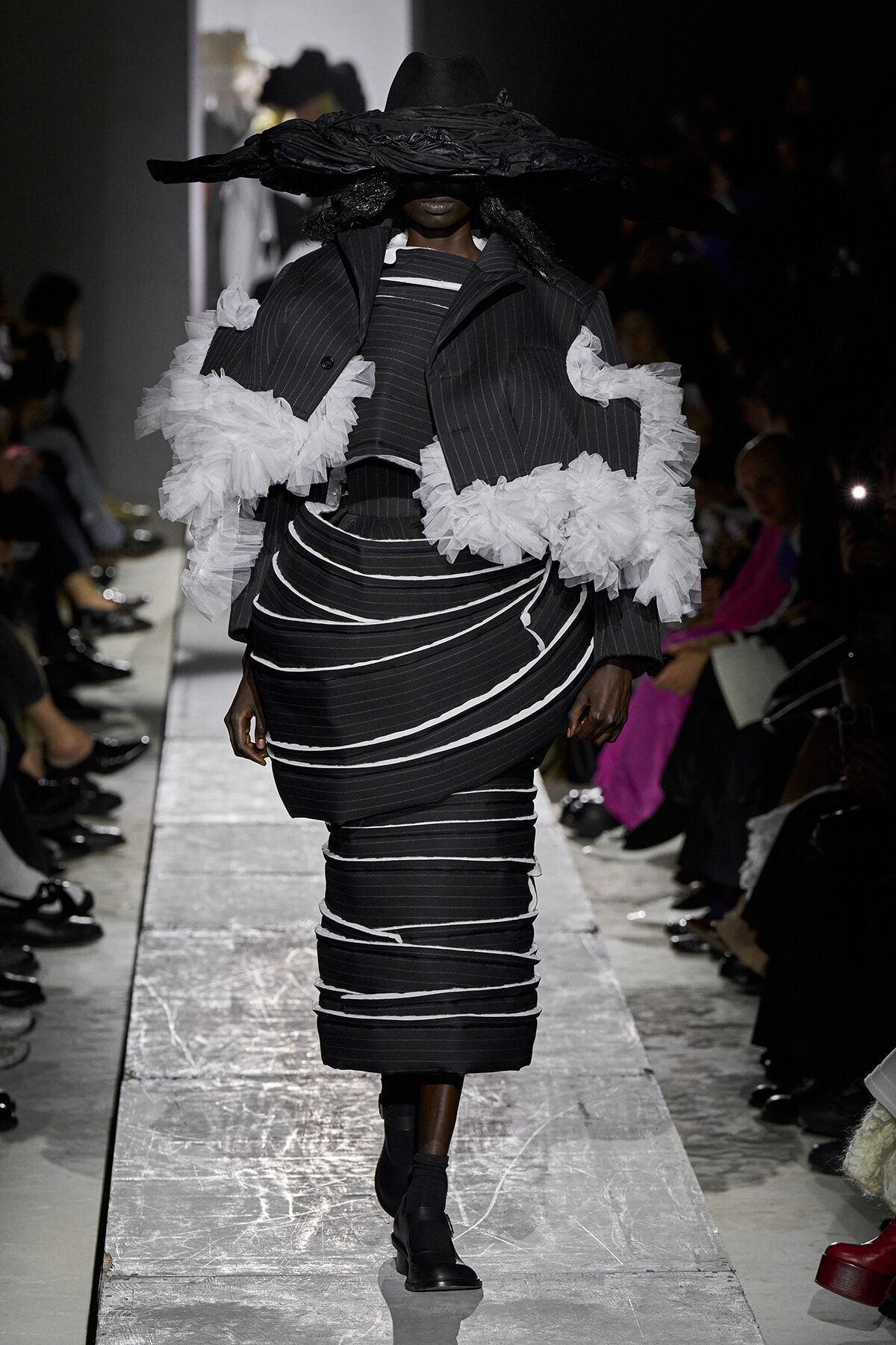 Runway model in a black pinstripe sculptural outfit with white ruffle sleeves and a wide-brimmed hat obscuring the face, black ankle boots visible.