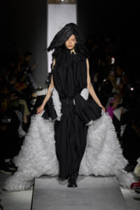Model walks runway in a black sculptural gown with a voluminous white ruffled train.
