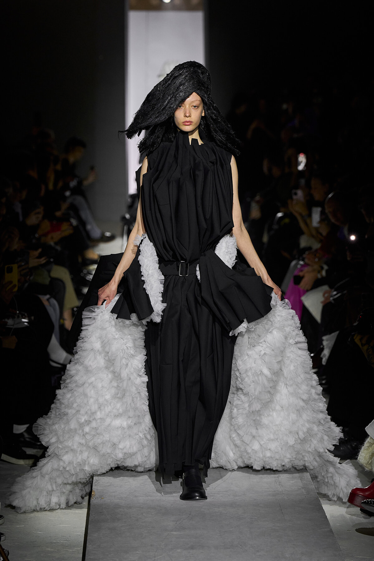 Model walks runway in a black sculptural gown with a voluminous white ruffled train.