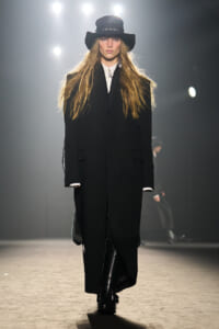 Model walks a runway in a long black coat and wide-brim black hat under bright stage lights.