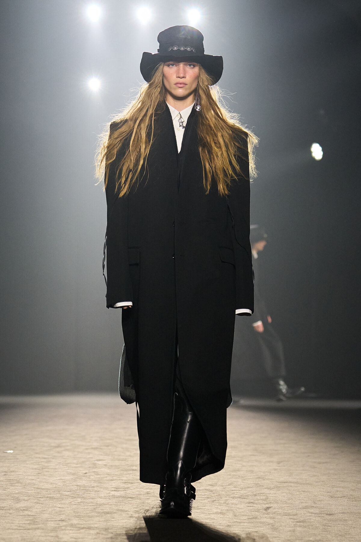 Model walks a runway in a long black coat and wide-brim black hat under bright stage lights.