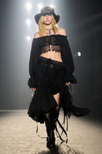 Model walks the runway in a black gothic-inspired outfit: off-the-shoulder lace crop top, layered skirt, and tall boots with a wide-brim hat.