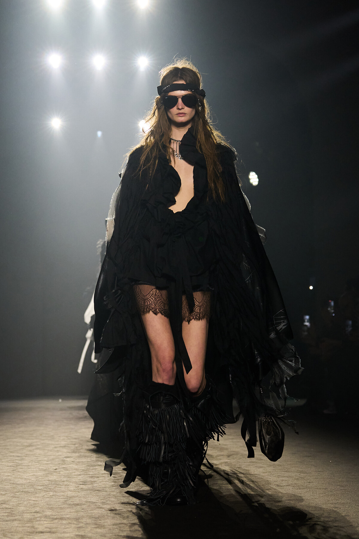 Model on a runway in an all-black look: flowing cape, plunging top, lace stockings, and fringed boots with sunglasses under bright stage lights.