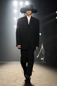 Runway model in an oversized black suit with white shirt, wide-brim black hat, walking under bright stage lights.