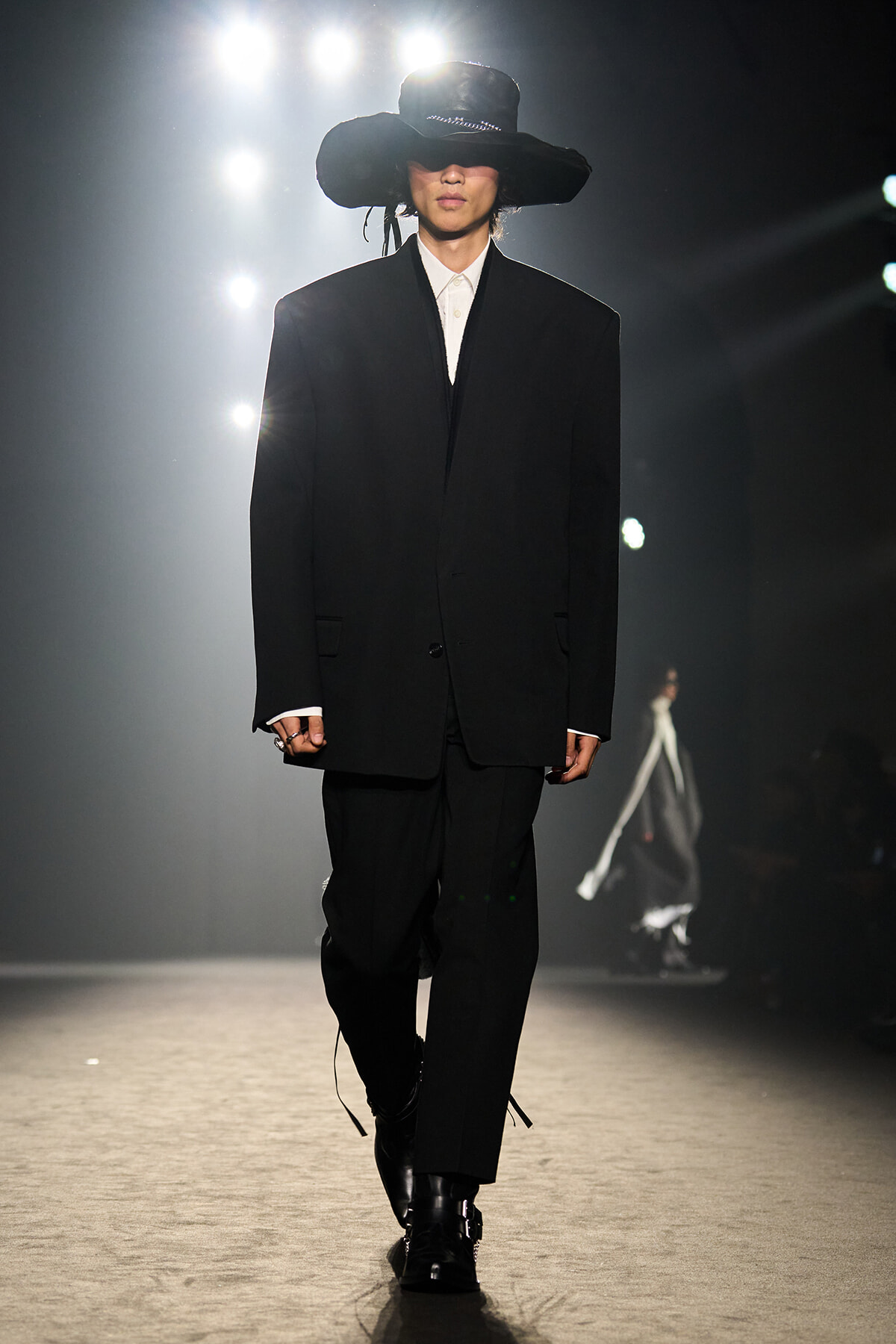 Runway model in an oversized black suit with white shirt, wide-brim black hat, walking under bright stage lights.