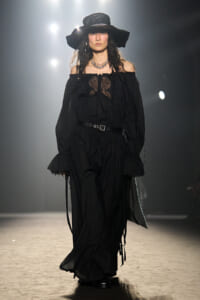 Model walking a runway in an all-black off-shoulder dress with a wide-brim hat and layered jewelry.