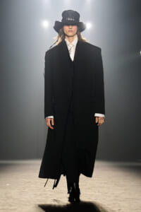 Model on a fashion runway wearing a long black coat, white shirt, and oversized black hat under bright spotlights.