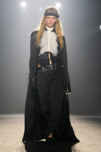 Model on a runway wearing a black Victorian-inspired ensemble with a white feathered bib, wide belt, and lace-up trousers on the catwalk.