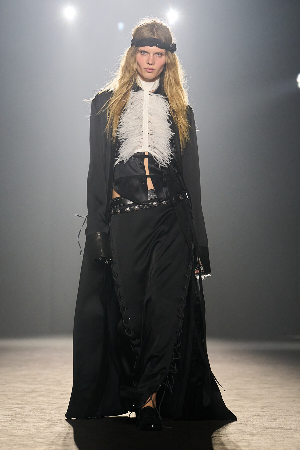 Model on a runway wearing a black Victorian-inspired ensemble with a white feathered bib, wide belt, and lace-up trousers on the catwalk.